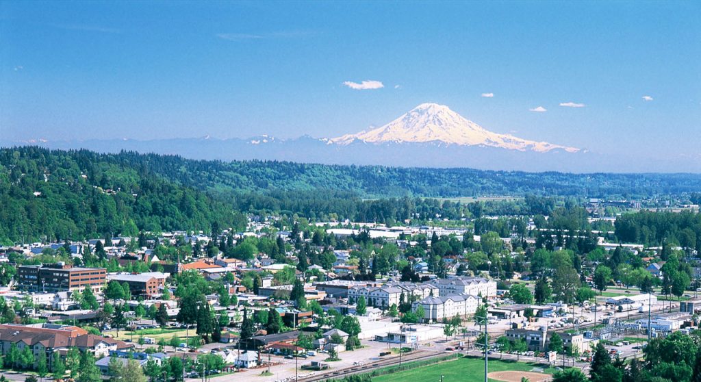 A city with Mount Rainier in the background, green hills, and a clear blue sky.