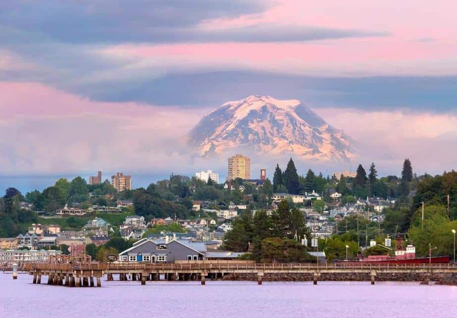 Snow-capped mountain looms over a city with a pier at sunset.